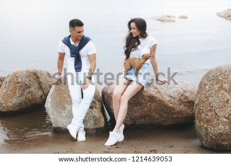 Young beautiful positive couple with small dog sitting on stone by sea. Lifestyle and pets