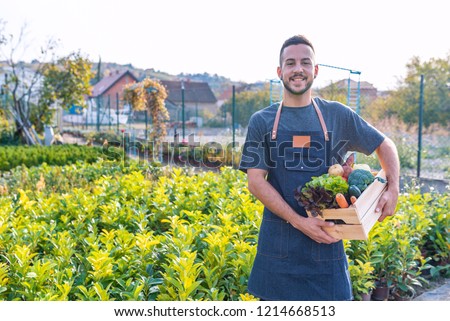 Smiling young farmer wearing straw hat and jean jacket, and looking at camera. Farmer holding wooden box with fresh vegetables and standing in nice green garden