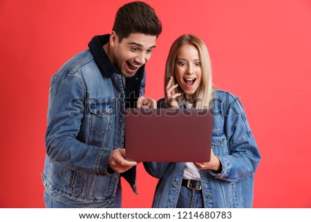 Portrait of an excited young couple dressed in denim jackets standing together isolated over red background, looking at laptop computer