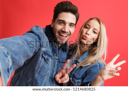 Portrait of a smiling young couple dressed in denim jackets standing together isolated over red background, taking a selfie, showing peace gesture