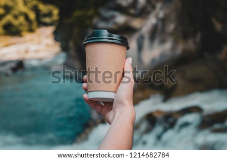 layout paper cups for beverages with a takeaway (tea or coffee) which holds female hand on the background of nature, against mountains and mountain river with forest. Coffee with you in a tourist spot