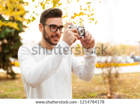 Young handsome hipster guy walks in a beautiful autumn park on the background of yellow leaves in warm sunny weather and takes pictures on a pig film camera. Autumn leisure time. Creative youth.