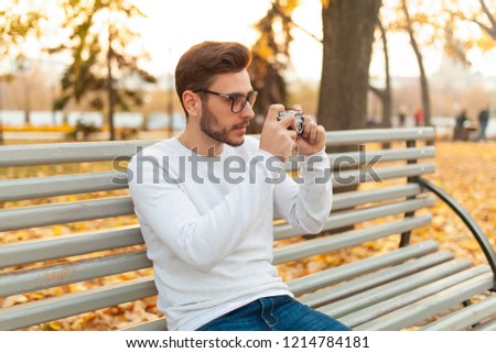 Young handsome hipster guy walks in a beautiful autumn park on the background of yellow leaves in warm sunny weather and takes pictures on a pig film camera. Autumn leisure time. Creative youth.