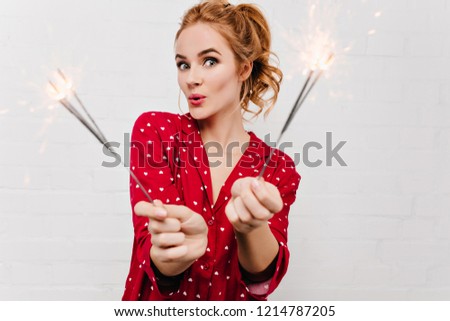Surprised woman in red pajama posing with sparkler. Cute blonde lady in funny night-suit holding bengal lights in christmas morning.