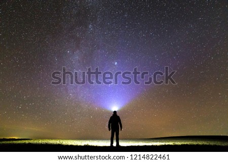 Back view of man with head flashlight standing on green grassy field under beautiful dark blue summer starry sky. Night photography, beauty of nature concept. Wide panorama, copy space background.