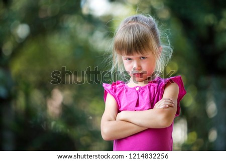 Portrait of pretty funny moody young blond child girl in pink sleeveless dress looks in camera feeling angry and unsatisfied on blurred summer green copy space background. Children tantrum concept.