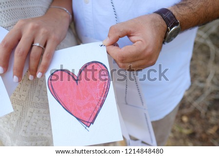 Close-up portrait of male and female hands holding bright red paper heart protecting it together. Love concept. Selective focus. Blurred background
