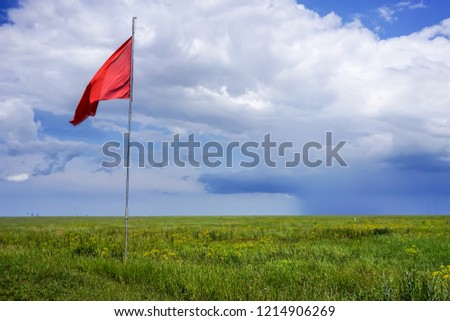 The steppe is covered with bright green grass with a flagpole and red flag against the blue sky and white clouds