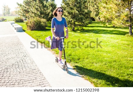 Woman riding a scooter. The girl in a dress rides a scooter in the park. Brunette pushes off the road. Feet in sneakers.