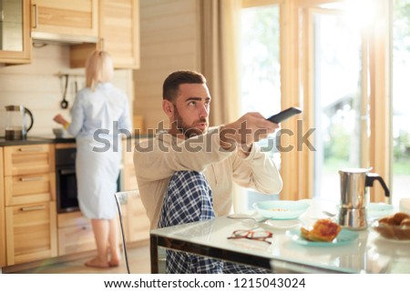 Young bearded man sitting at kitchen table and switching channels on TV with remote control while his wife making breakfast