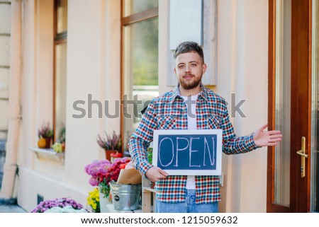 Cheerful, nice man owner floral store in informal wear holding opem sign, having his hands open like hospitable gesture, you are welcome, well met, standing over storefront of flower shop, outdoor.
