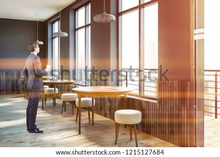Businessman standing in corner of modern cafe with gray walls, wooden floor, round wooden tables and small round chairs along the window. Toned image