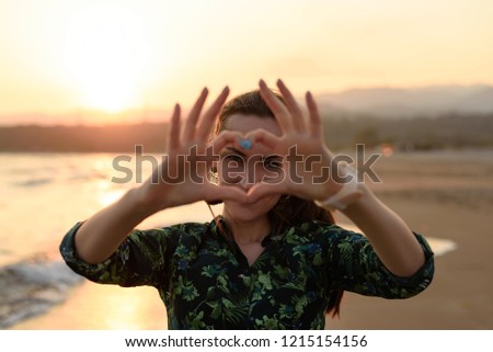 Portrait of a young woman on the beach at red sunset, heart of fingers, message of love.