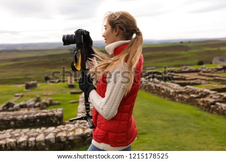 Girl photographer standing near the ancient ruins. Wearing  the red waistcoat and gloves. Trying to take good pictures of the nature. Green field around her. Forest on the background. 