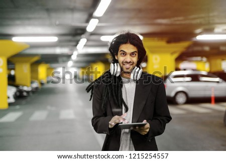 Young cute urban hipster man holding tablet and looking at camera. Standing in garage with headphones around his neck.