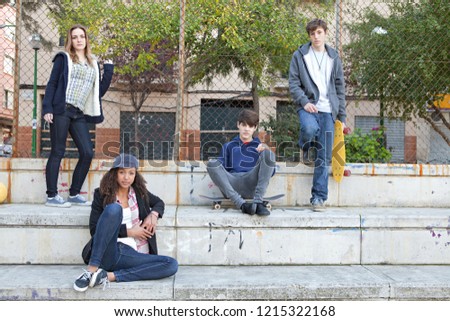 Group of four ethnic diverse teenagers relaxing together in sports court with skateboards, looking trendy outdoors. Healthy adolescents, leisure recreation lifestyle.