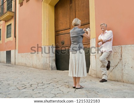 Mature couple visiting old city street monument with stone pavement holding smartphone taking photos, holiday outdoors. Senior man and woman using technology, leisure recreation lifestyle.