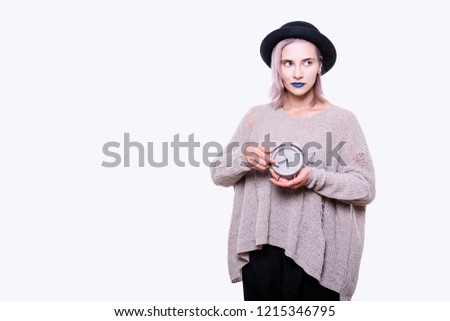 Portrait of young emotional attractive woman in black hat, torn sweater, with blue lips and pink hair, she is holding grey alarm clock, isolated on white background. Time management concept