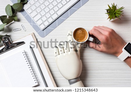 cropped image of businessman with prosthesis hand and smartwatch sitting at table with coffee cup and computer keyboard in office