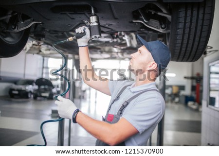 Picture of younf man standing and working underneath car. He looks up. Guy holds and uses drill. He works in white gloves.