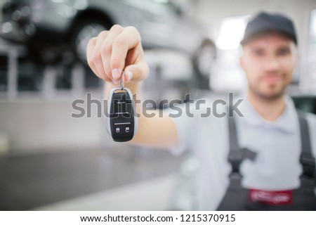 Blurred picture of key that worker hold in hand. He look at it and smile a bit. Young man stand in garage. There is car on platform behind him.