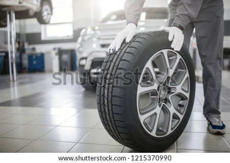 Picture of worker stand and hold both hands on tire. He wears gloves. Guy poses. Car stands behind him.