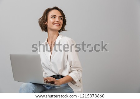 Image of young business woman posing isolated over grey wall background sitting on stool using laptop computer.