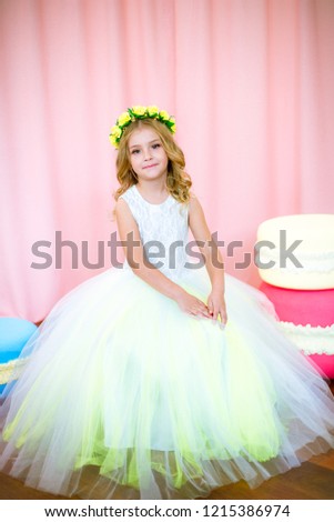 Cute little girl with blond curls in a white lush dress with giant sweets in pink background studio