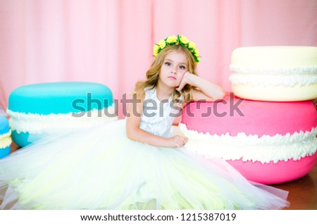 Cute little girl with blond curls in a white lush dress with giant sweets in pink background studio