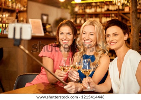 people, technology and lifestyle concept - women drinking wine and taking picture by smartphone on selfie stick at bar or restaurant
