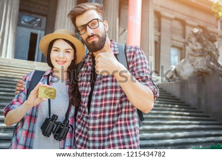 Couple of young tourists stand outisde at stairs and pose on camera. She holds gold card in hand. Man hold big thumb up. She has binoculars on her chest.