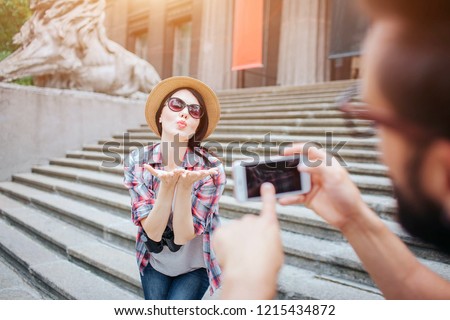 Beautiful young woman poses on camera and sending kisses to young bearded man. She stands on stairs. Guy holds phone in hands. He points on screen.