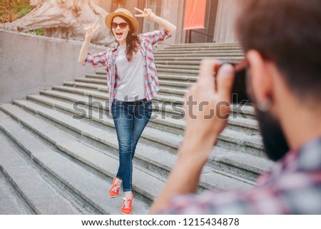 Young attractive woman stands on stairs and poses on camera. She keeps hands up and wears glasses with hat. Female touris is excited. Yong bearded man takes pictures of her.