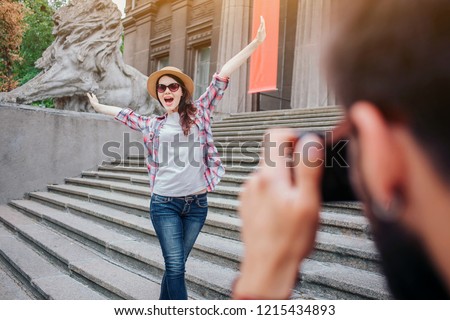 Young bearded man talking picture of happy woman. She poses on camera. Female tourist keep hands up and wears glasses. They stand on stairs.