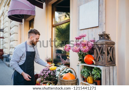 Friendly handsome worker in apron decorating facade store with orange pumpkin and chrysanthemums outdoor over showcase of floral,decor,gift shop. Flowers delivery, creating order, small business. Royalty-Free Stock Photo #1215441271