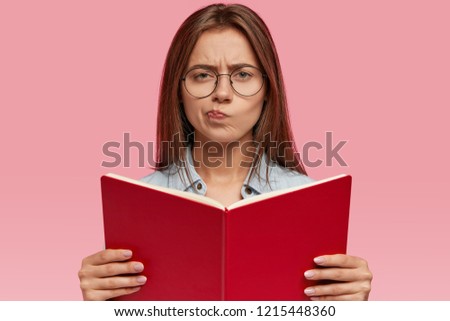 People, education concept. Puzzled beautiful woman with long dark hair, holds red book, wears round spectacles, has displeased expression as feels bored of reading, isolated over pink background