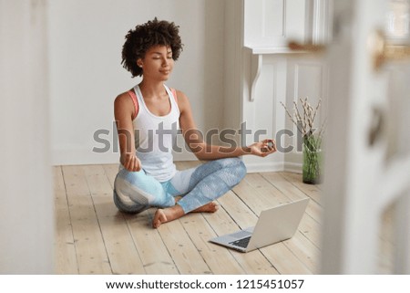 Relaxed dark skinned woman with sporty body, sits in zen pose, keeps legs crossed, watches yoga lessons on laptop computer using internet, meditates on floor in empty room. Wellness concept. Royalty-Free Stock Photo #1215451057