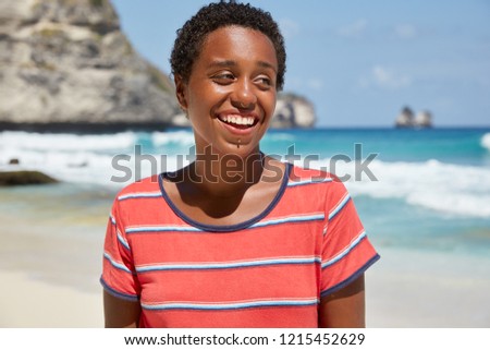 Panoramic view of black girl hipster has broad smile, piercing in nose and near mouth, boycut, dressed in casual striped t shirt, poses against azure ocean view and rocky mountains. Bright blue colors