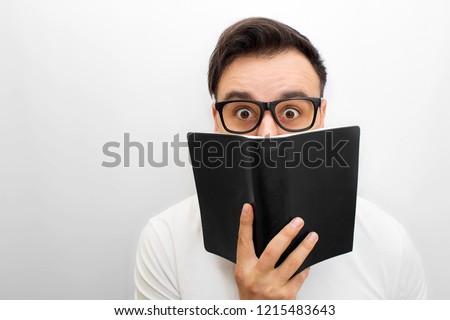 Picture of male brunette looking on camera. He holds black book in hands. He weas glasses. Young man is excited. Isolated on white backgorund.