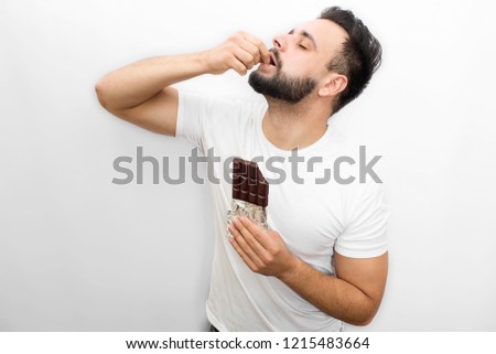 Picture full of pleasure. Young bearded man stands and eats chocolate. He puts piece of it into mouth. Guy keeps eyes closed. Man is concentrated. Isolated on white background.