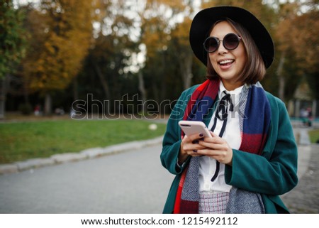 Cheerful and stylish hipster girl in hat using smart phone and  laughing. Portrait of modern girl having smart phone in hands using 4G internet.