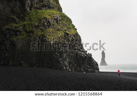 Woman in red raincoat taking pictures of the black basalt columns at the black sand beach in Vik, Iceland.