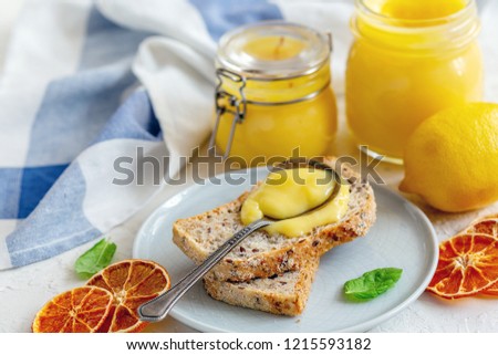 Slice of whole grain bread with lemon custard on a plate and glass jars with lemon custard on a textured white table, selective focus.