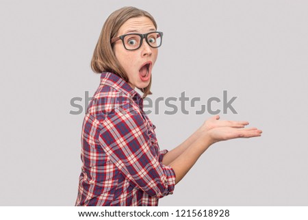 Exited and amazed young woman poses on camera. She holds two hands together. Model keeps mouth wide opened. She wears glasses. Isolated on grey background.