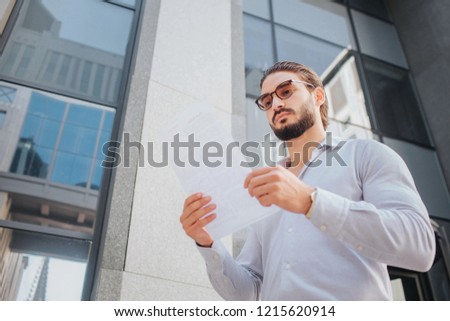 Picture of young stylish man stand and poses. He looks at white piece of paper through sunglasses. Guy is calm, peaceful and concentrated. He stands in front af glass building.
