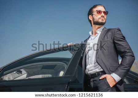 Picture of well-dressed businessman stand at black car with its door opened. He holds one and in pocket and another one on car door. Young man poses.