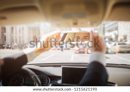 Picture of man's eyes looking straight from rare view mirror. He holds it with both hands. Guy sits in car, He has suit on himself.