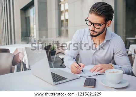 Picture of young bsinessman works at table. He writes down in opened notebook and look at laptop's screen. Guy is calm and concnetrated. There is white cup at table.