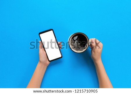Female hands holding black mobile phone with blank white screen and mug of coffee. Mockup image with copy space. Top view on blue background, flat lay.