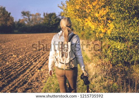 Woman holding a camera and walking on footpath in countryside at autumn. Beautiful colorful autumn rural scenery with female photographer